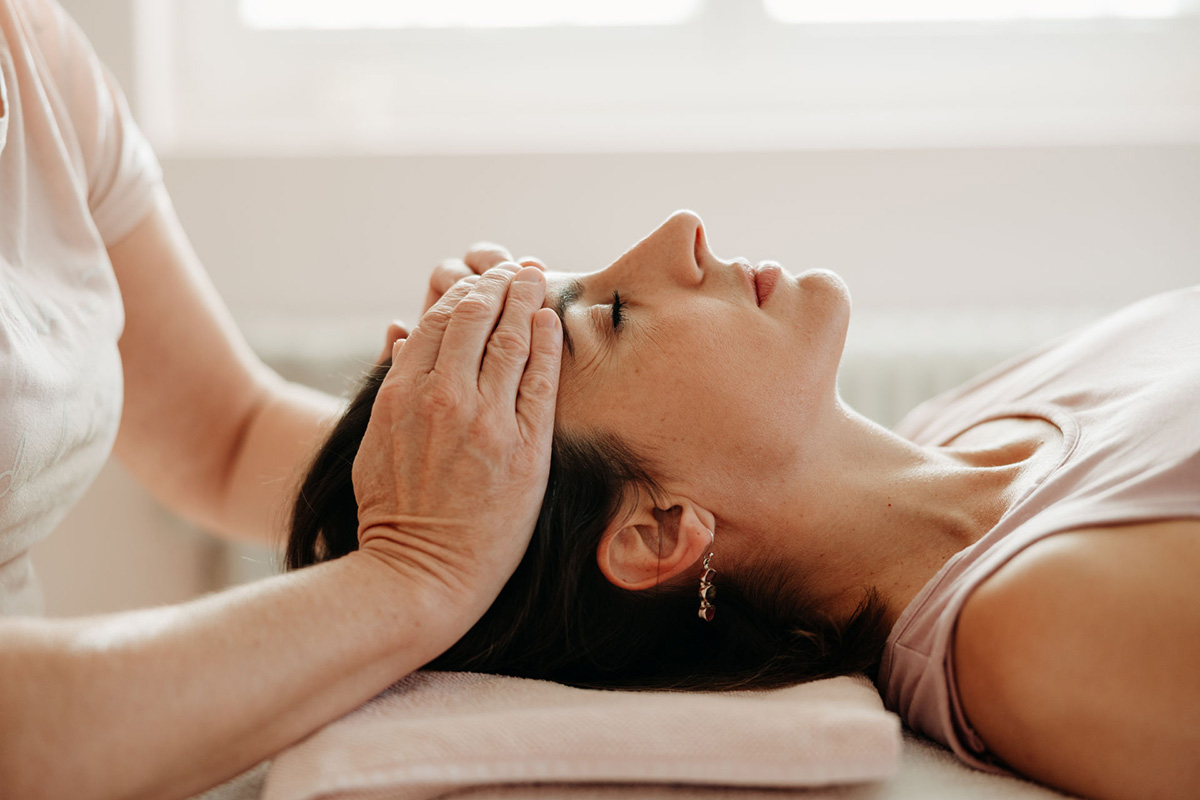 Eine Frau, liegend, mit geschlossenen Augen. Zwei Hände liegen auf Ihrer Stirn