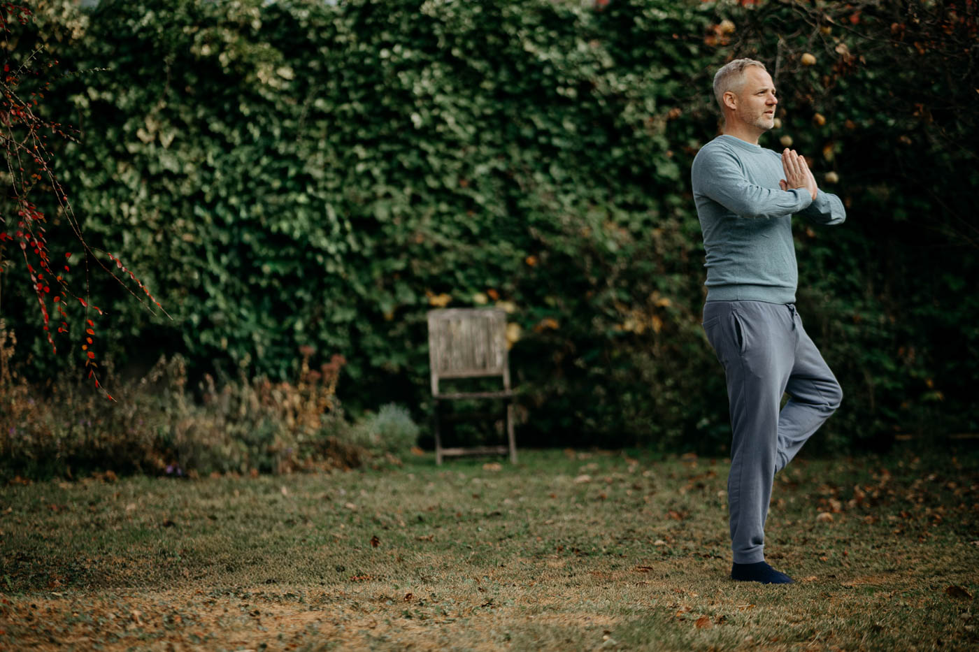 Ein Mann in Yogapose steht auf einer Wiese, im Hintergrund sind Büsche