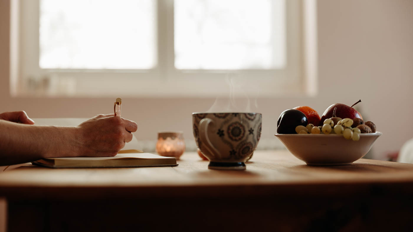 Ein Tisch mit einr Teetasse, einer Früchteschale und einer Hand, die einen Stift hält und in ein Buch schreibt